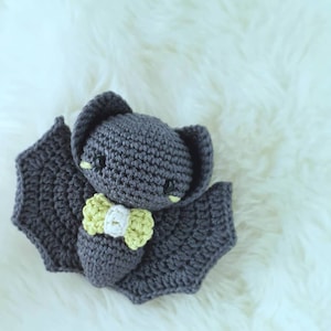 May include: A gray crocheted bat with yellow and white bow tie. The bat is sitting on a white fluffy surface.