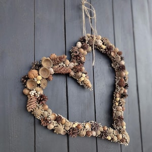May include: A heart-shaped wreath made of natural materials, including pinecones, acorns, and berries. The wreath is hung on a dark gray wooden door.