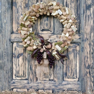 May include: A rustic wreath made of wine corks, burlap, and grape clusters hangs on a weathered wooden door. The wreath is decorated with dried leaves and vines.