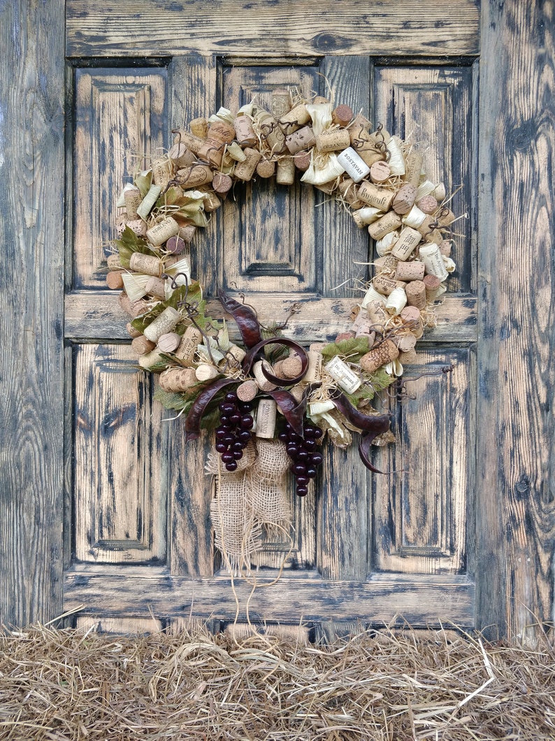 May include: A rustic wreath made of wine corks, burlap, and grape clusters hangs on a weathered wooden door. The wreath is decorated with dried leaves and vines.