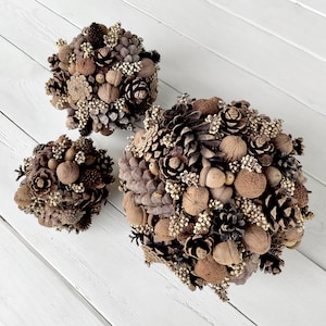 May include: Three brown, round, decorative arrangements made of pinecones, acorns, and other natural elements. The arrangements are on a white wooden surface.