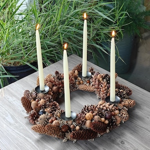 May include: An Advent wreath with four lit ivory candles. The wreath is made of pinecones, nuts, and berries, set on a dark grey metal base. Green plants form the background, creating a warm, natural aesthetic.