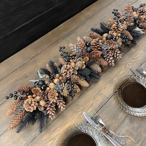 May include: A decorative table runner featuring a mix of pinecones, berries, and dark green and black foliage. The arrangement is set on a wooden table, with clear glass plates and silverware on either side.
