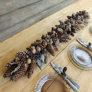 May include: A table centrepiece made from pine cones, nuts, and foliage. The arrangement is in shades of brown and tan, with varied textures. Clear glass plates and cutlery are set on a light wood table, creating a rustic, natural aesthetic.