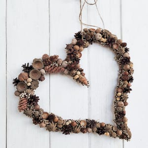 May include: A heart-shaped wreath made of natural materials, including acorns, pinecones, and berries. The wreath is brown and beige and hangs on a white wooden door.