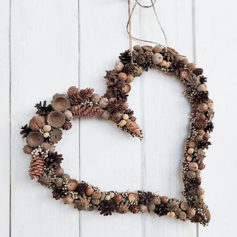 May include: A heart-shaped wreath made of natural materials, including acorns, pinecones, and berries. The wreath is brown and beige and hangs on a white wooden door.