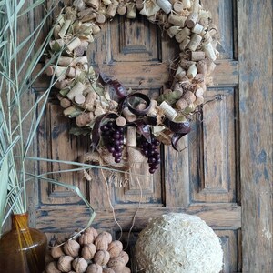 May include: A rustic wreath made of wine corks, burlap, and grape vines. The wreath is decorated with faux grapes and is hanging on a wooden door. There are three decorative balls made of natural materials on a wooden table in front of the door.