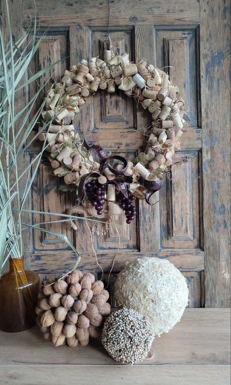 May include: A rustic wreath made of wine corks, burlap, and grape vines. The wreath is decorated with faux grapes and is hanging on a wooden door. There are three decorative balls made of natural materials on a wooden table in front of the door.