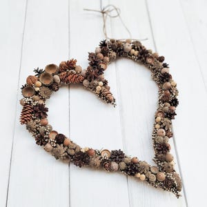 May include: A heart-shaped wreath made of natural elements, including pinecones, acorns, and dried flowers. The wreath is brown and beige in color and is hanging on a white wooden background.