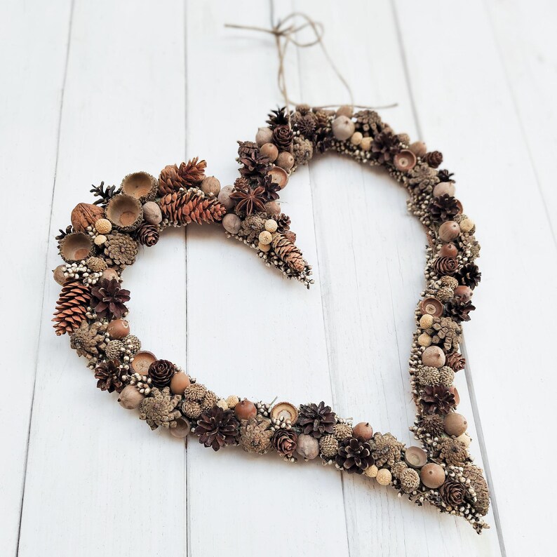May include: A heart-shaped wreath made of natural elements, including pinecones, acorns, and dried flowers. The wreath is brown and beige in color and is hanging on a white wooden background.