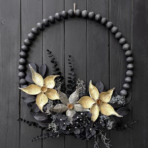 May include: A circular wreath constructed from matte black wooden beads, adorned with two large gold-toned dried flowers, a central grey textured seed pod, dark black foliage, and delicate wisps of light grey dried botanical accents.