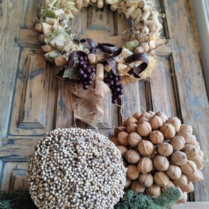 May include: A rustic wreath made of wine corks, burlap, and grapes hangs on a wooden door. Two decorative spheres, one made of small white beads and the other of walnuts, sit on a wooden surface in front of the door.