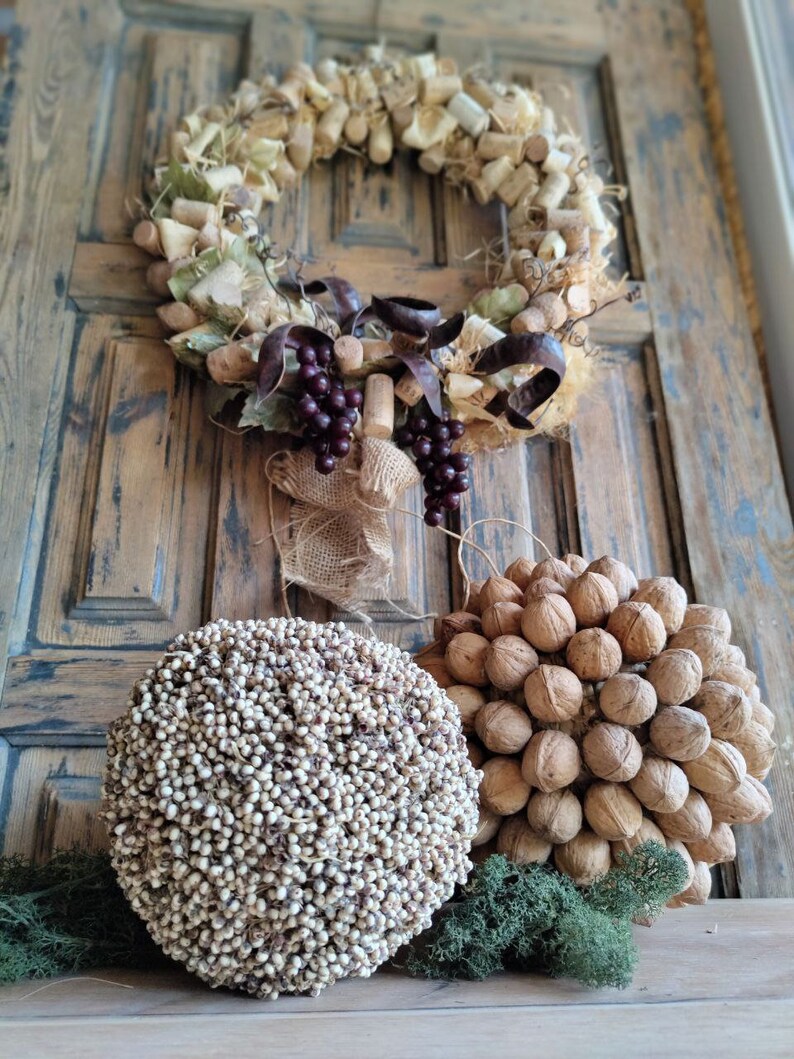 May include: A rustic wreath made of wine corks, burlap, and grapes hangs on a wooden door. Two decorative spheres, one made of small white beads and the other of walnuts, sit on a wooden surface in front of the door.