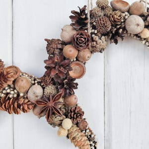 May include: A brown and beige wreath made of natural elements, including pinecones, acorns, and dried flowers. The wreath is shaped like a heart and is perfect for a rustic or farmhouse decor style.