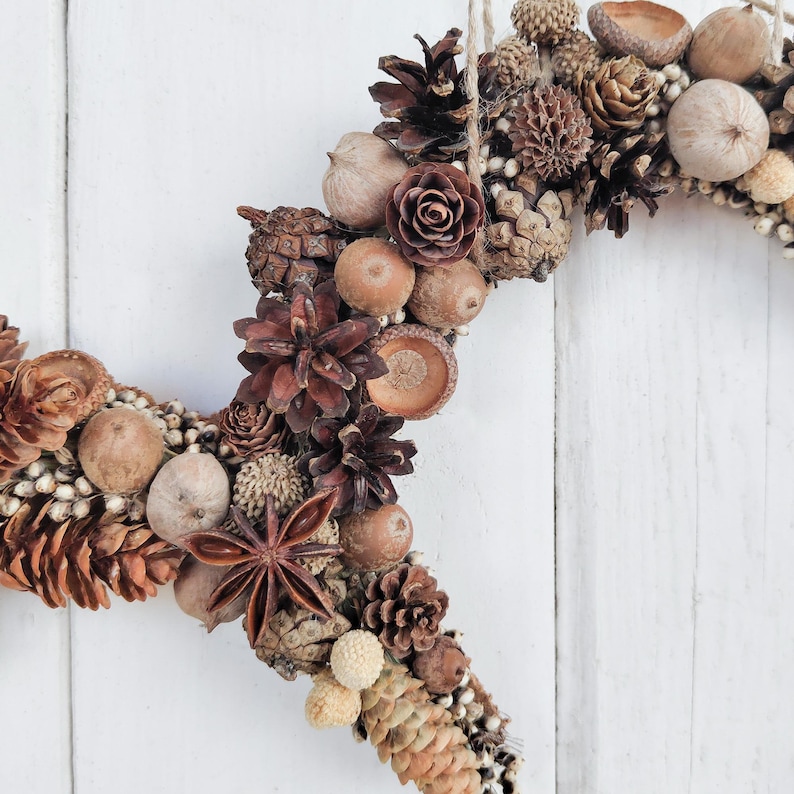 May include: A brown and beige wreath made of natural elements, including pinecones, acorns, and dried flowers. The wreath is shaped like a heart and is perfect for a rustic or farmhouse decor style.