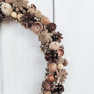 May include: A brown, natural, autumnal wreath made of pinecones, acorns, and dried flowers. The wreath is hanging on a white wooden background.