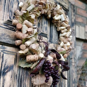 May include: A rustic wreath made of wine corks, burlap, and grape clusters. The wreath is decorated with dried leaves and is hanging on a wooden door.