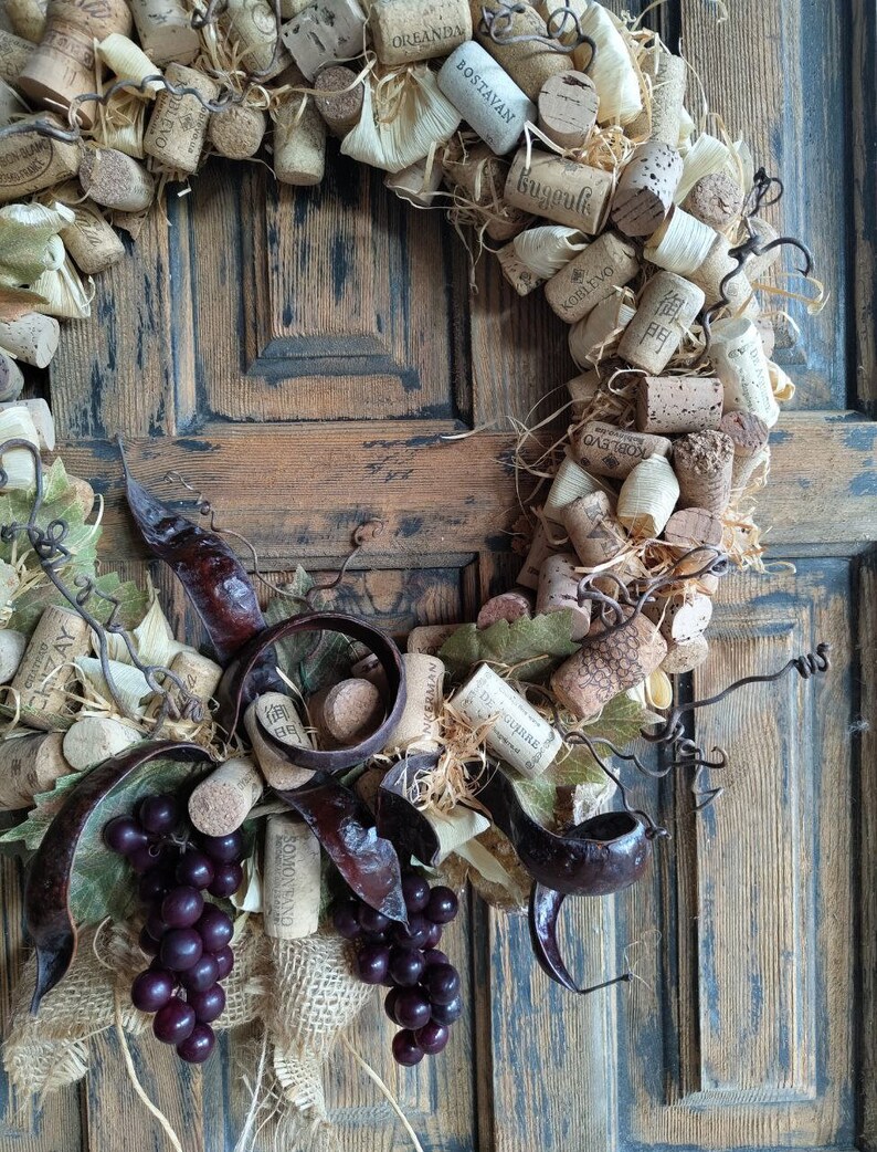 May include: A rustic wine cork wreath with faux grapes and burlap accents. The wreath is decorated with wine corks with various labels, including "OREANDA", "BOSTAVAN", "KOBLEVO", and "BUGULMA".