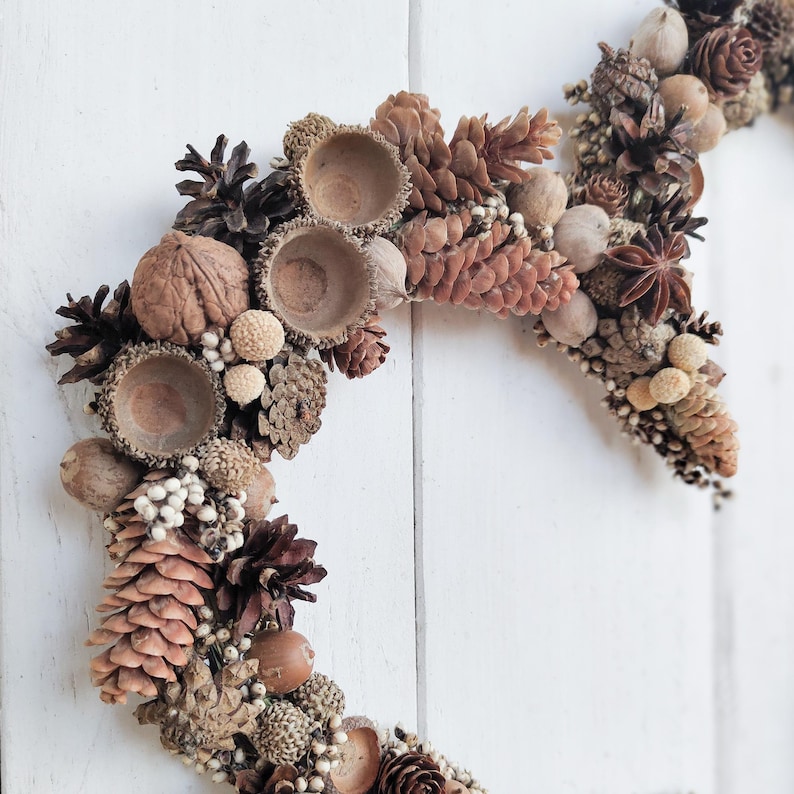 May include: A brown, natural, autumnal wreath made of pinecones, acorns, and other natural elements. The wreath is in the shape of a crescent moon.