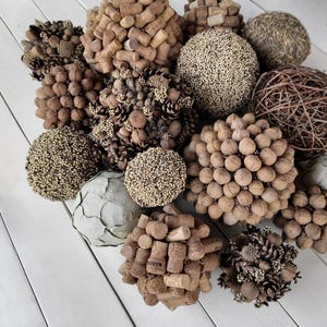 May include: A collection of decorative spheres made from various natural materials, including cork, walnuts, pine cones, and twigs. The spheres are arranged on a white wooden surface.