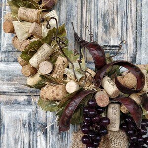 May include: A grapevine wreath decorated with wine corks, dried leaves, and burlap ribbon. The wreath is adorned with a cluster of dark purple grapes and features the text "SOMONTANO" on a wine cork.