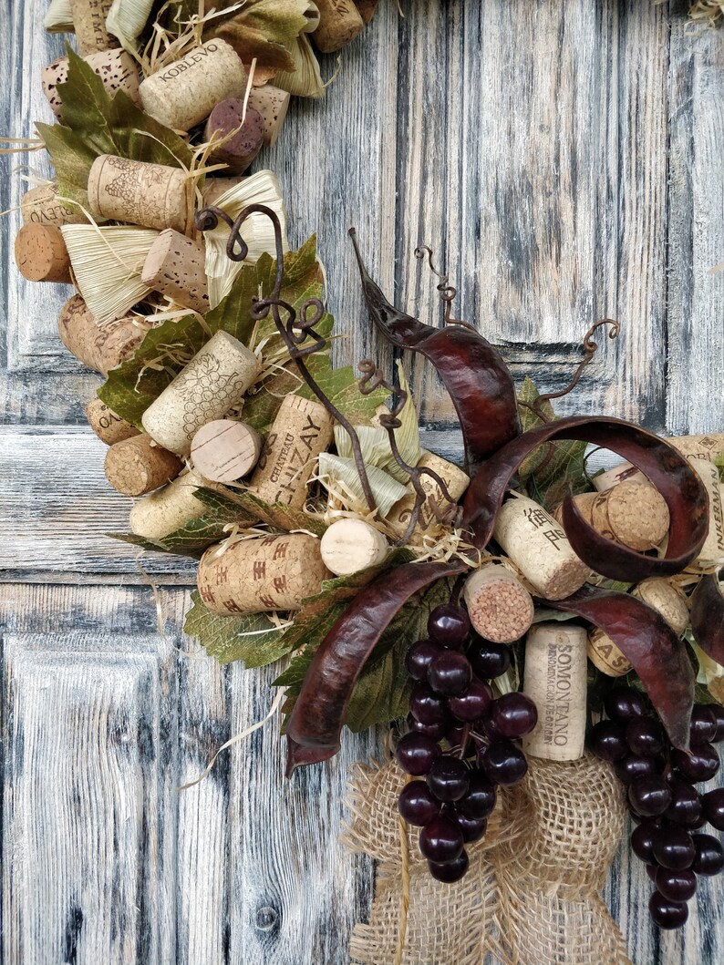 May include: A grapevine wreath decorated with wine corks, dried leaves, and burlap ribbon. The wreath is adorned with a cluster of dark purple grapes and features the text "SOMONTANO" on a wine cork.