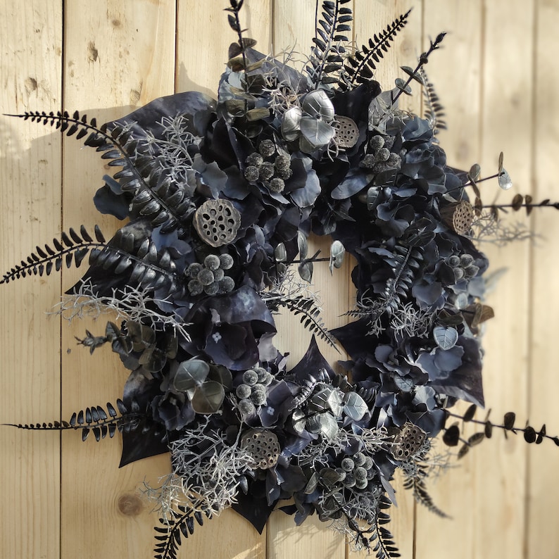 May include: A black and gray wreath made of dried flowers, leaves, and pods. The wreath is decorated with black berries and has a rustic, natural look.