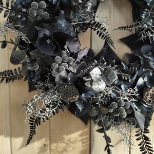 May include: A black and silver wreath with eucalyptus leaves, berries, and lotus pods. The wreath is decorated with silver accents and has a rustic, vintage feel.