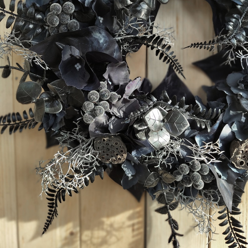 May include: A black and silver wreath with eucalyptus leaves, berries, and lotus pods. The wreath is decorated with silver accents and has a rustic, vintage feel.