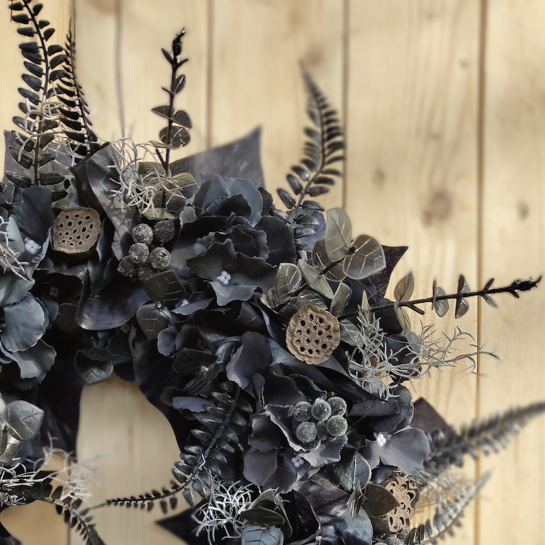 May include: A black and gray wreath with hydrangeas, eucalyptus leaves, and dried lotus pods. The wreath is hanging on a wooden wall.