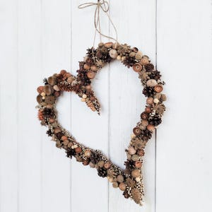 May include: A heart-shaped wreath made of natural materials, such as pinecones, acorns, and berries. The wreath is brown and beige and is hanging on a white wooden wall.