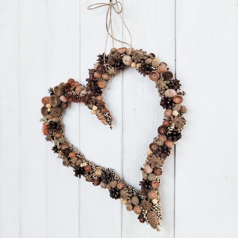 May include: A heart-shaped wreath made of natural materials, such as pinecones, acorns, and berries. The wreath is brown and beige and is hanging on a white wooden wall.