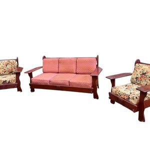 May include: A three-piece wooden living room set. The set includes a sofa and two armchairs. The sofa and armchairs have reddish-brown wooden frames and floral patterned cushions. The cushions have a cream background with orange, yellow, and brown flowers.