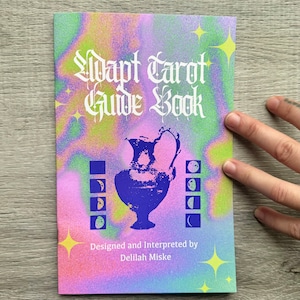 Op de afbeelding: Een "Adapt Tarot Guide Book" met een kleurrijk, gespikkeld ontwerp. De cover toont een blauwe vaasillustratie, maanfase-afbeeldingen en de tekst "Adapt Tarot Guide Book" in een gotisch lettertype. Ontworpen en geïnterpreteerd door Delilah Miske.