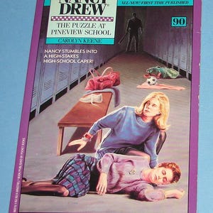 May include: A purple and white book cover with the title "Nancy Drew" and the subtitle "The Puzzle at Pineview School". The cover features an illustration of two girls in a school changing room, one lying on the floor and the other standing over her.