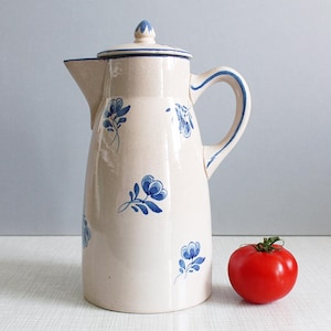 以下が含まれることがあります： クリーム色のセラミックピッチャー。青い花の模様と青いハンドルが付いています。熟した赤いトマトがピッチャーの隣にあります。ピッチャーの高さは約25cmです。