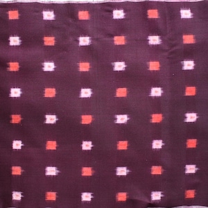 以下が含まれることがあります： 濃い紫色の背景に、赤とピンクの正方形が繰り返されるパターンの生地。生地は質感があり、端が処理されています。