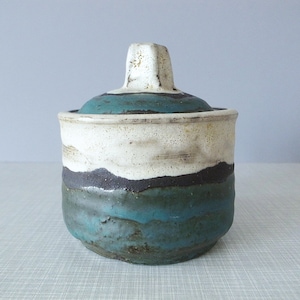 以下が含まれることがあります： 白、黒、青の釉薬が施された蓋付きの陶器の壺。壺は素朴で手作り感のある外観です。