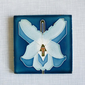 Può includere: Una piastrella di ceramica blu con un design di orchidea bianca. L'orchidea è stilizzata e ha una forma geometrica. La piastrella ha una finitura lucida.