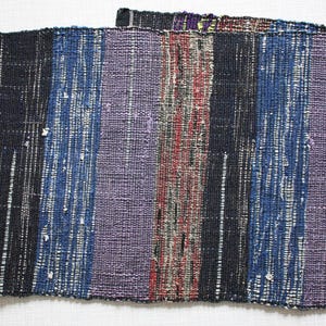 May include: A woven textile with vertical stripes in shades of purple, blue, black, red, and brown. The fabric appears to be handwoven, with a textured surface. The textile could be used as a table runner or decorative accent.