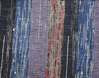 May include: A woven textile with vertical stripes in shades of purple, blue, black, red, and brown. The fabric appears to be handwoven, with a textured surface. The textile could be used as a table runner or decorative accent.