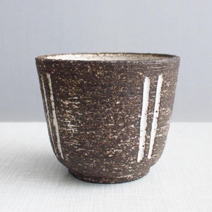 以下が含まれることがあります： 白い縦線が引かれた茶色の陶器のカップ。カップはざらざらとした質感の表面をしています。