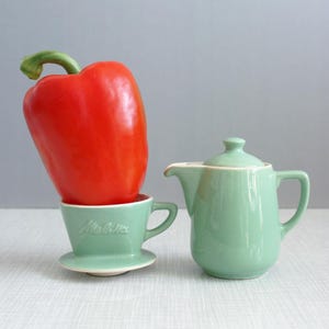 May include: A red bell pepper sits on top of a green ceramic coffee filter with the word "Melitta" printed on it. A green ceramic creamer sits next to the filter.