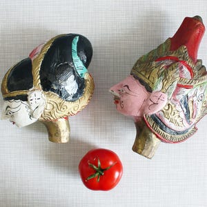 Puede incluir: Dos cabezas de marionetas de madera tallada, con rostros pintados y elaborados tocados. Una es negra y dorada, la otra roja y dorada. Entre ellas, un tomate rojo fresco, que añade un toque de color a la composición.