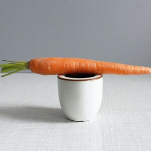 以下が含まれることがあります： 白いセラミック製の卵カップの縁に、緑の葉のついたオレンジ色のニンジンが1本置かれています。ニンジンは少し曲がっていて、カップは白いテクスチャのある表面の上に置かれています。