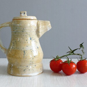 以下が含まれることがあります： 薄い黄色の釉薬と青い斑点のある陶器のティーポット。ティーポットには蓋と取っ手があります。背景には、つるに赤いトマトが3つあります。
