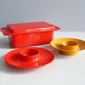 以下が含まれることがあります： 赤い蓋付きバターディッシュ、赤いソーサー付きエッグカップ、黄色いソーサー付きエッグカップの3つのヴィンテージキッチン用品