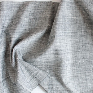 Puede incluir: Un trozo de tela gris con un tejido texturizado, parcialmente enrollado y drapeado. La tela tiene un patrón sutil y un borde sin rematar, lo que sugiere que puede usarse para proyectos de costura o artesanía. El color es un gris neutro.