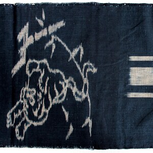 May include: A rectangular piece of dark navy blue fabric featuring a woven ikat pattern. The left side displays a stylized animal figure in light gray, while the right side shows a series of horizontal stripes and abstract gray shapes.