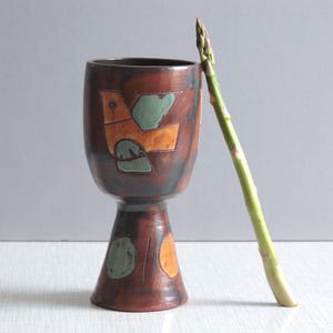 Puede incluir: Una copa de cerámica marrón con un motivo de pájaro pintado a mano en naranja y verde en el cuenco y formas circulares abstractas en la base, acompañada de un tallo de espárrago verde apoyado contra el lado del recipiente.